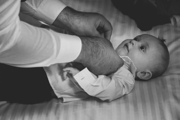 Father dressing his baby in formal attire for a wedding. The baby is lying down while the father adjusts the shirt. Captured in a black-and-white photograph by Chris Stephenson Photography, highlighting a tender, intimate moment.