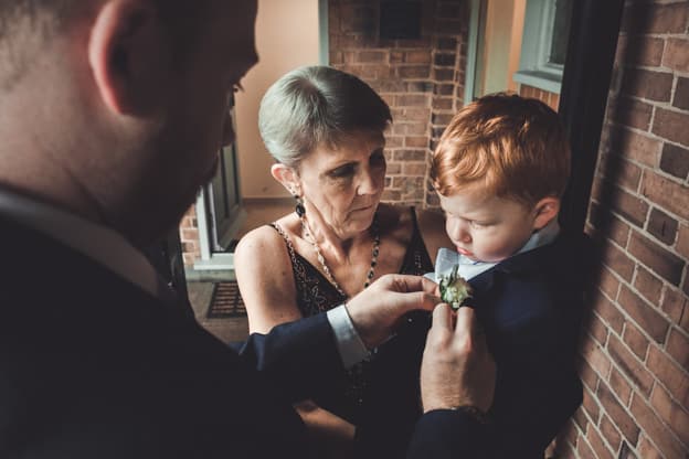 An older woman helping a young boy with his boutonnière while a man adjusts his suit. The boy, dressed in formal attire, stands patiently. Captured by Chris Stephenson Photography in a warm, candid moment.