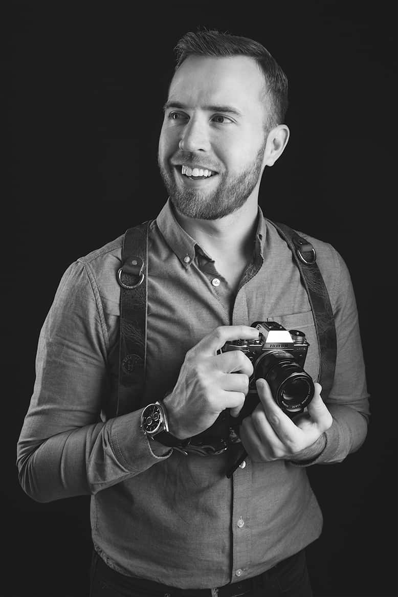 Chris Stephenson, a professional wedding photographer, smiling and holding a camera. He is dressed in a casual button-up shirt with a camera harness, ready for a photoshoot. Captured in a black and white portrait with a dark background.