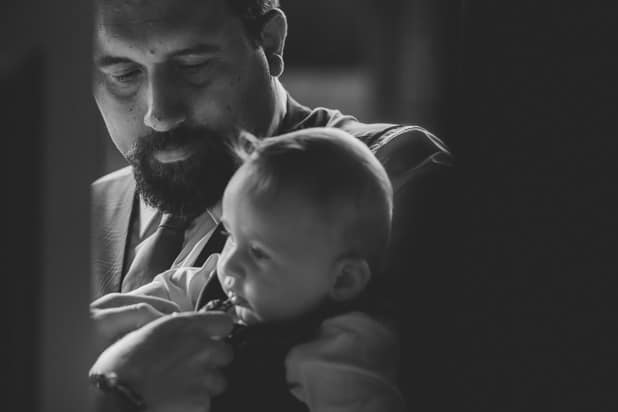 Father holding his young child during the wedding ceremony. The father has a beard and is dressed in a suit, while the child is dressed in formal attire. Captured in a tender, black-and-white moment by Chris Stephenson Photography.