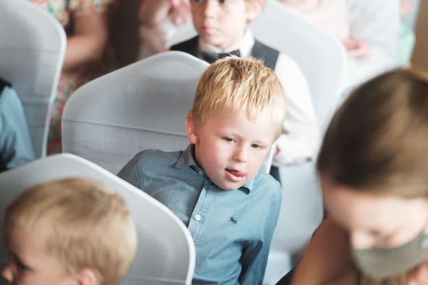 Young wedding guest sitting on a chair, looking attentively. The child is wearing a blue shirt and surrounded by other guests. Captured by Chris Stephenson Photography in a candid moment.