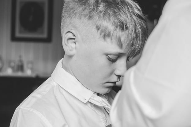 Young boy in a white shirt looking down with a thoughtful expression. Captured in a black-and-white photograph by Chris Stephenson Photography, highlighting a quiet, introspective moment during the wedding