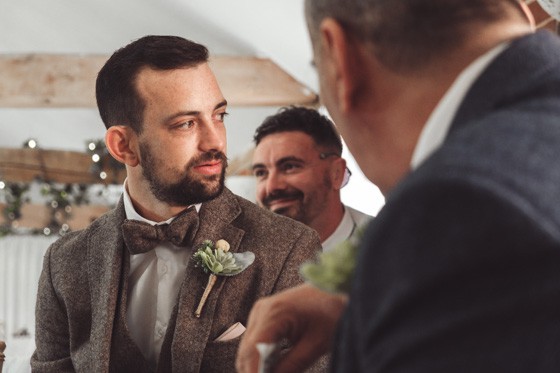A guest in a tweed suit with a bow tie, conversing with another guest at the wedding reception. Another guest is smiling in the background. Captured by Chris Stephenson Photography with a focus on candid, relaxed moments