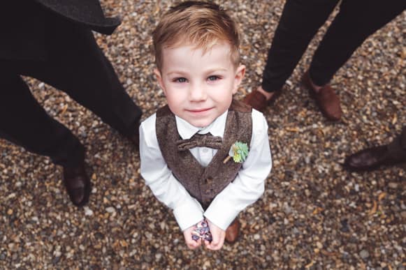 The bride's young son dressed in a tweed vest and bow tie, looking up and smiling. Captured by Chris Stephenson Photography in a charming, candid moment.