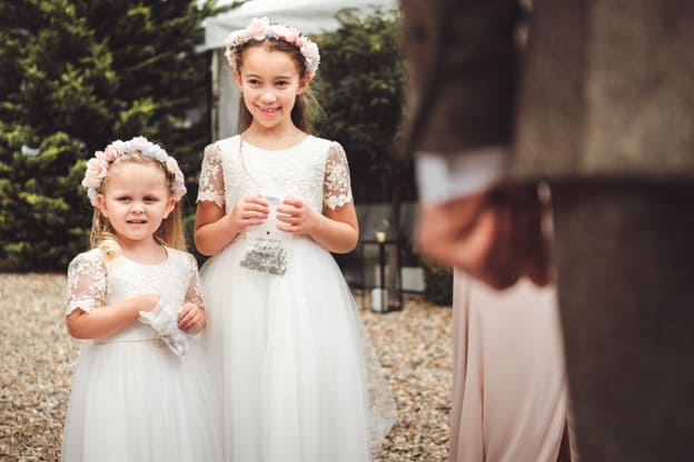 Two young flower girls dressed in white dresses with lace sleeves, holding small bags, and wearing flower crowns. They are smiling and standing outdoors, ready for the wedding ceremony. Captured by Chris Stephenson Photography in a charming, candid style.