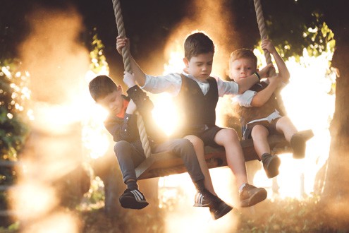 Three young boys sitting on a swing together, dressed in formal attire, with sunlight streaming through the trees in the background. Captured by Chris Stephenson Photography in a playful, candid moment.