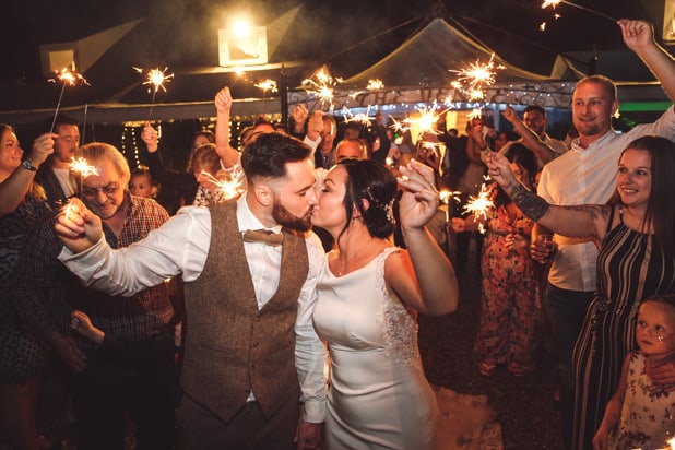 Bride and groom kissing during their wedding celebration, surrounded by guests holding sparklers at night. The bride is in a white dress and the groom in a brown vest and bow tie. Captured by Chris Stephenson Photography in a festive, candid style.